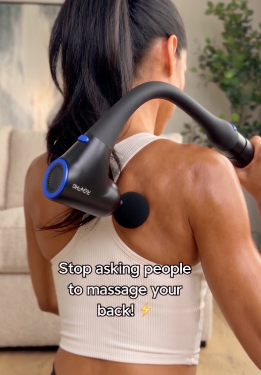 Woman using a self back massage gun with extension handle to reach upper back knots and relieve neck and shoulder pain at home